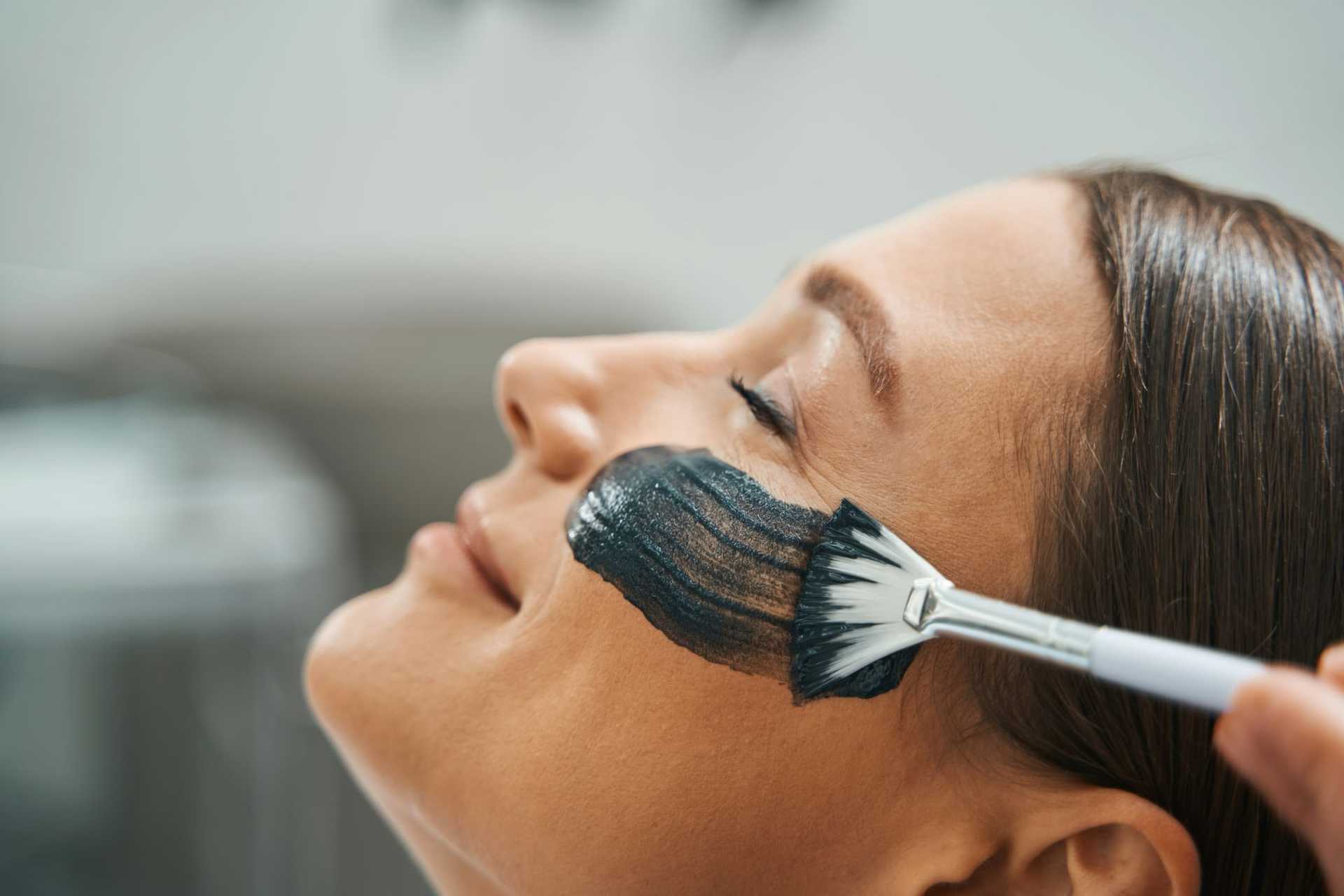 Woman receiving a black facial mask treatment with a brush at a spa.