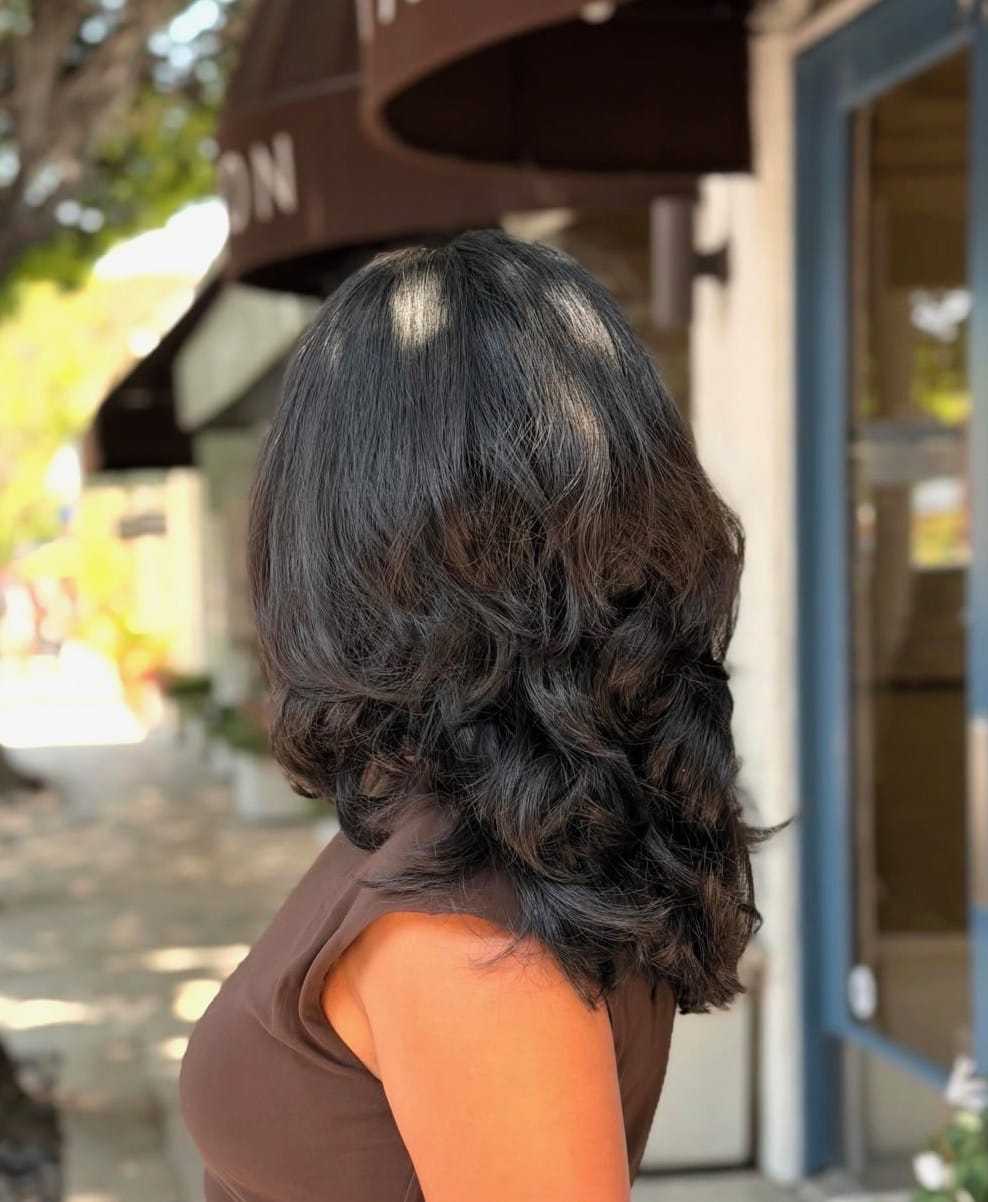 Back view of a woman with long, layered dark hair standing on a sunlit street.