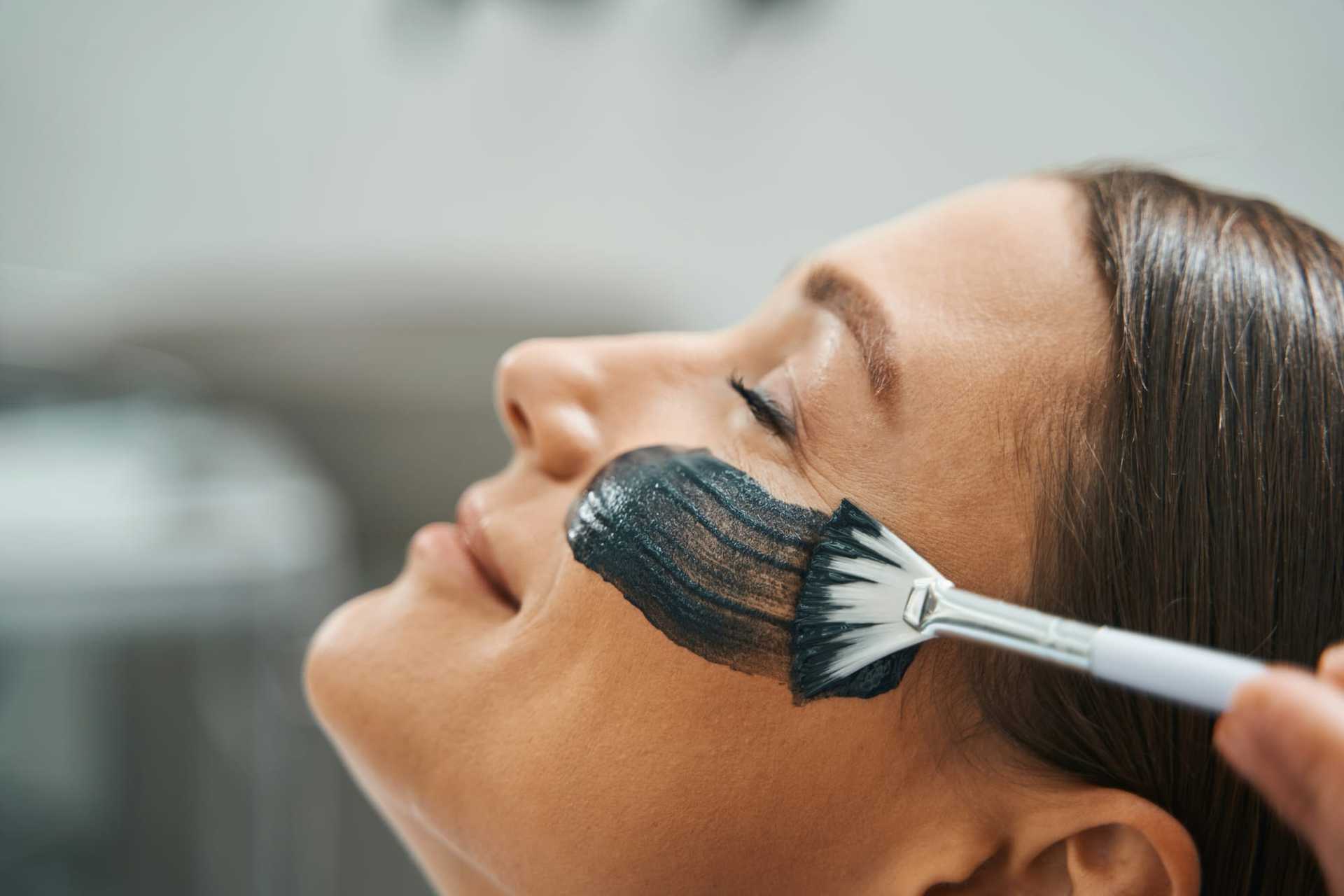 Woman receiving a black facial mask treatment with a brush at a spa.