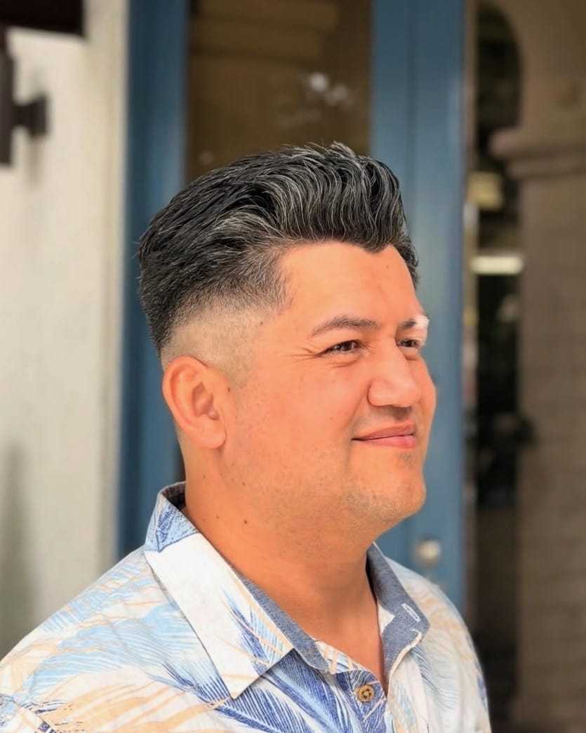 Man with styled hair smiling, wearing a patterned shirt, standing outside a building.