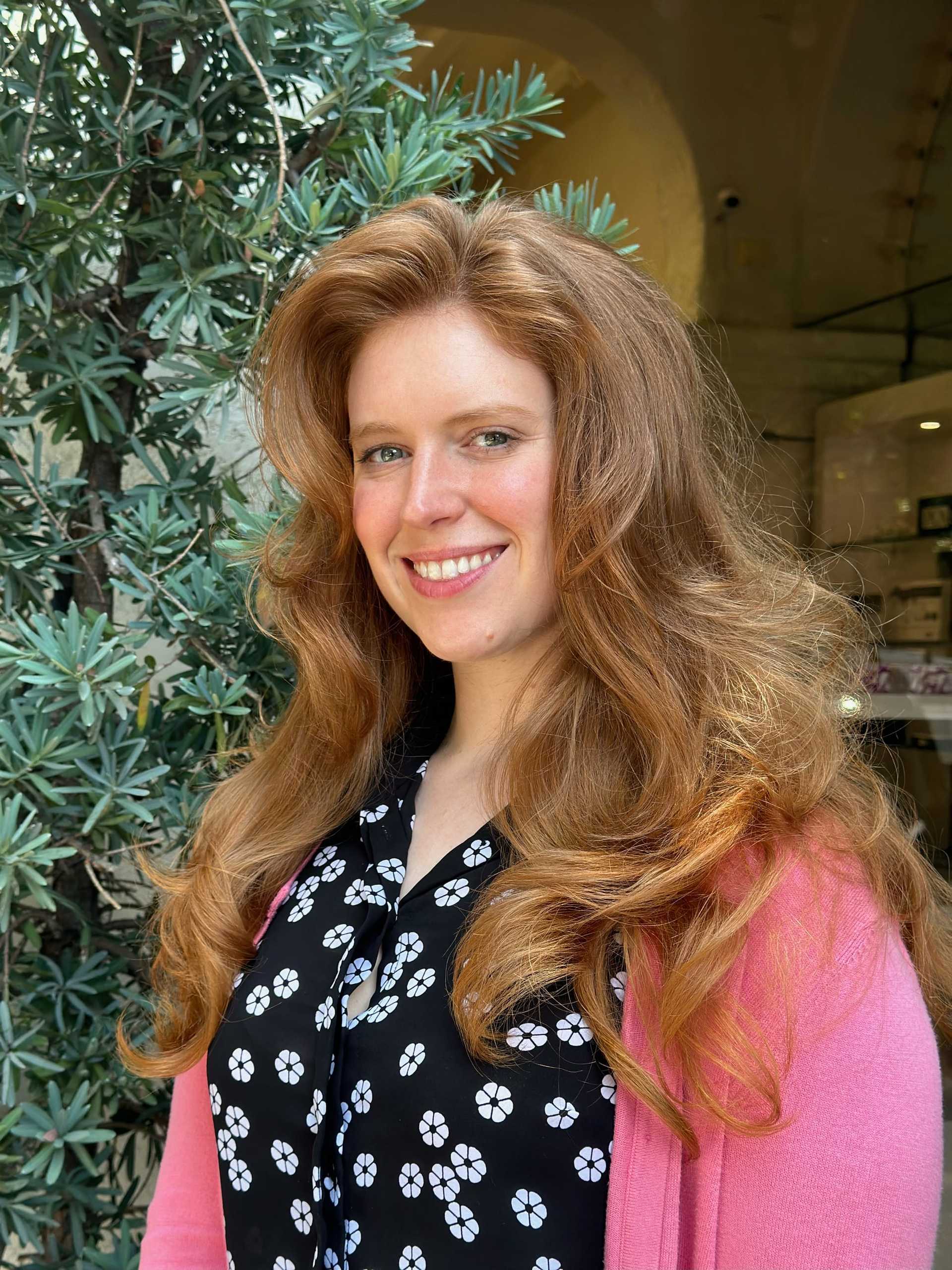 Smiling woman with long red hair stands by a leafy plant in a sunlit area.